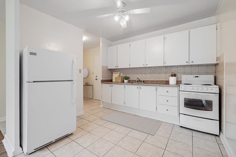 A white refrigerator stands in a kitchen with white cabinets and a white stove.